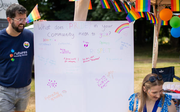 A male member of staff stands next to a canvas reading "What does Pride in your community mean to you", looking down towards a woman wearing blue and with brightly coloured face paint.