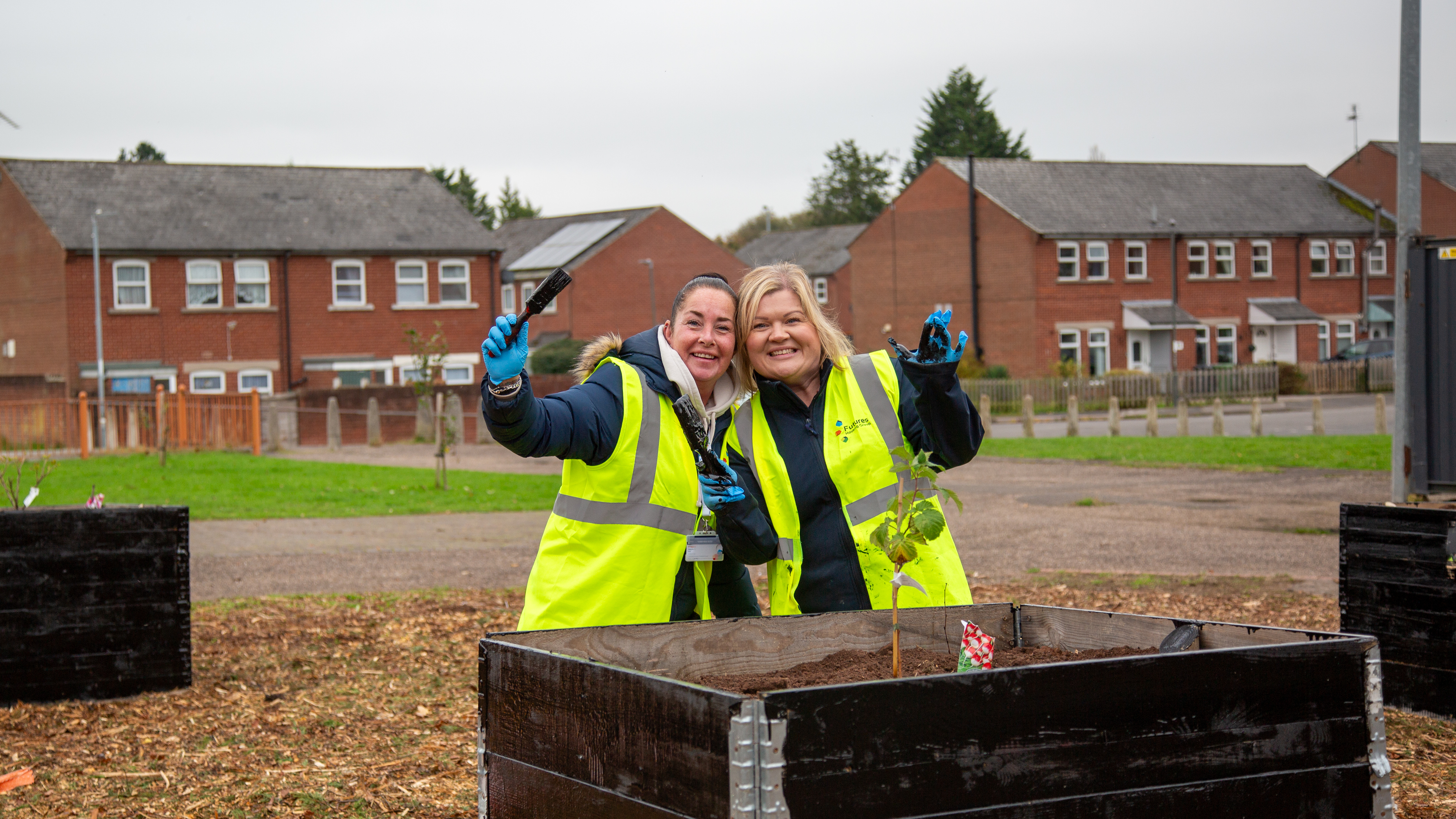 Two smiling people in hi-vis jackets posing for a picture holding paintbrushes in front of a planter