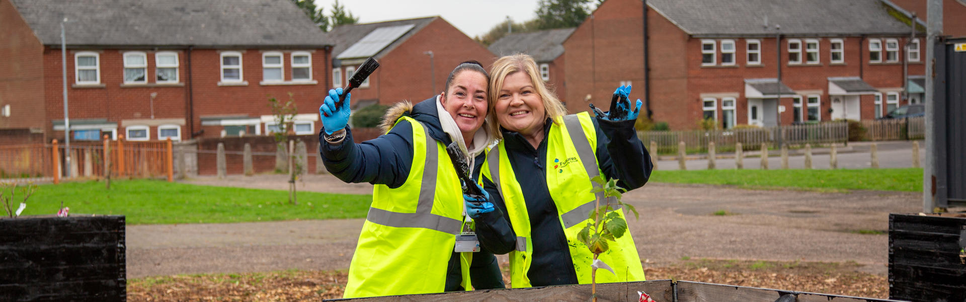 Two smiling people in hi-vis jackets posing for a picture holding paintbrushes in front of a planter