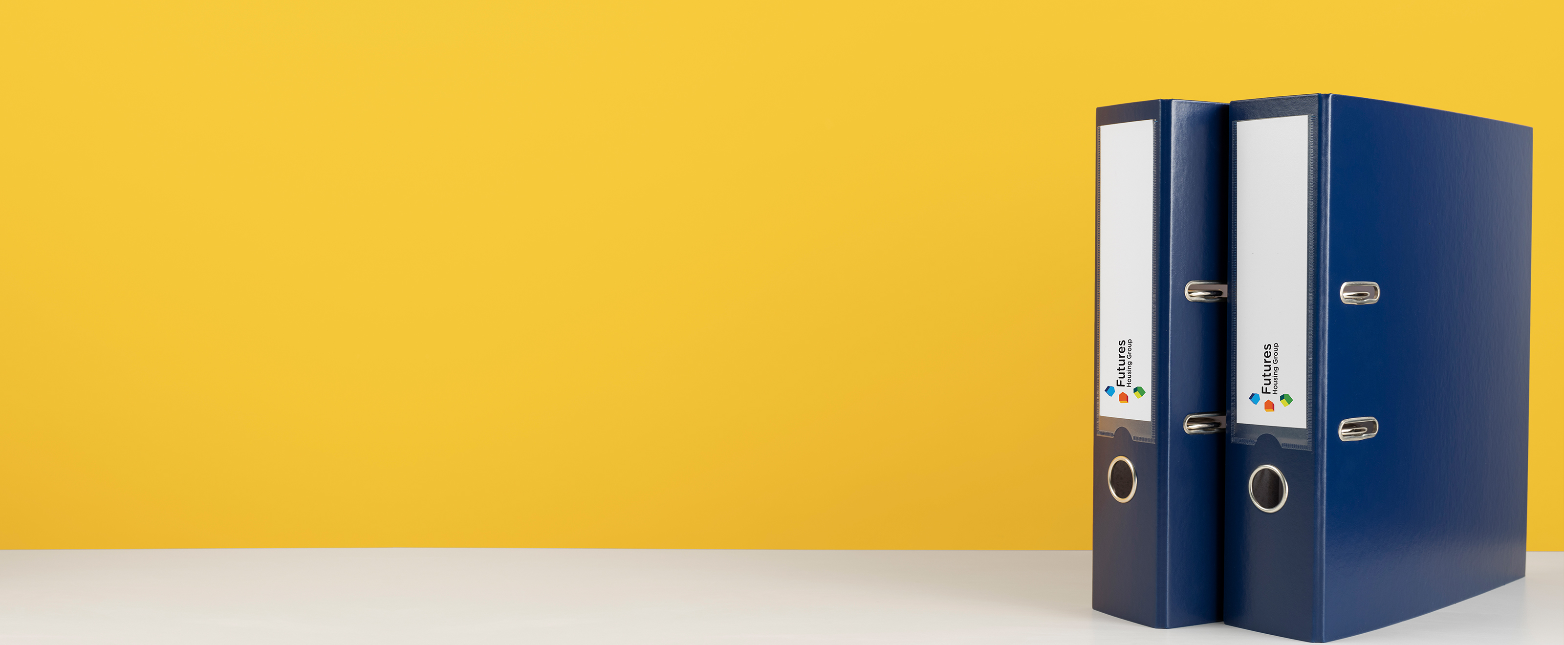 Photo of two blue binder files on a white surface with a yellow background. The spines of the files have a white area with the Futures Housing Group logo on it.