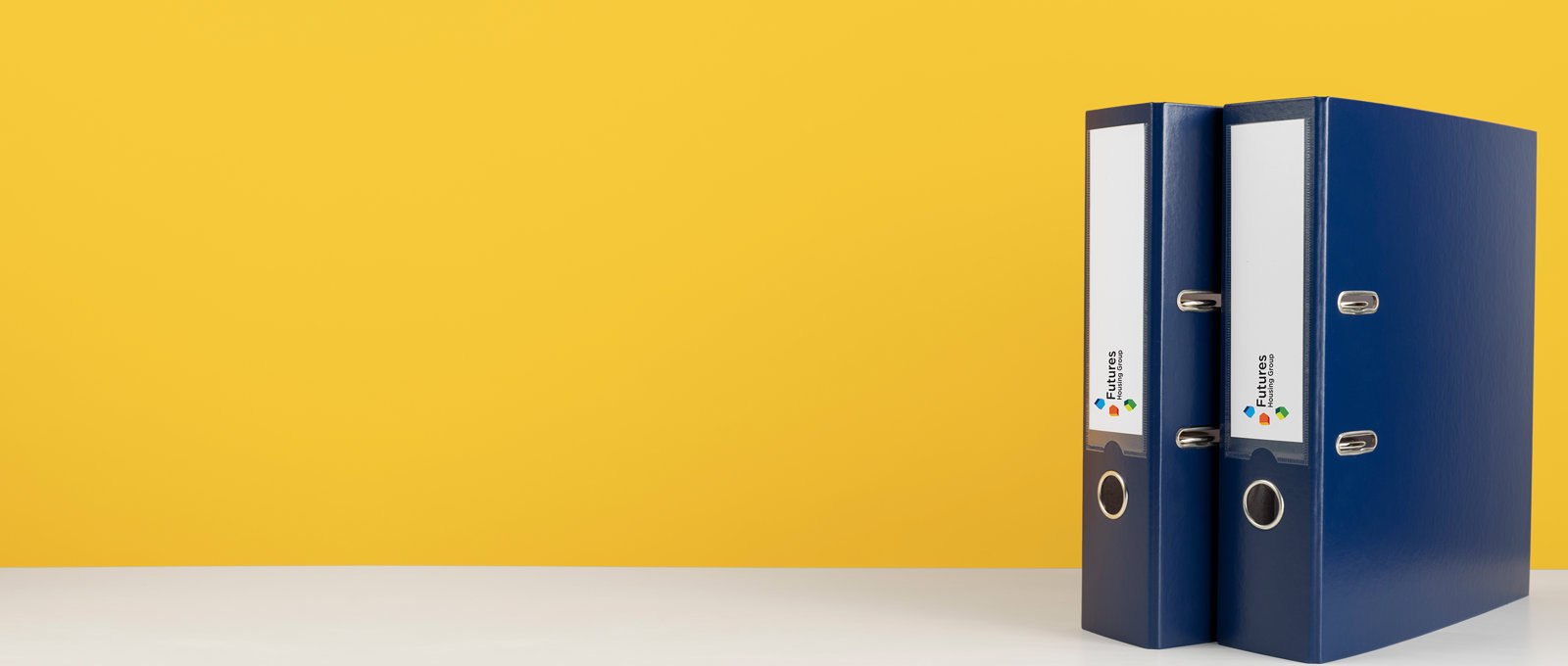 Photo of two blue binder files on a white surface with a yellow background. The spines of the files have a white area with the Futures Housing Group logo on it.