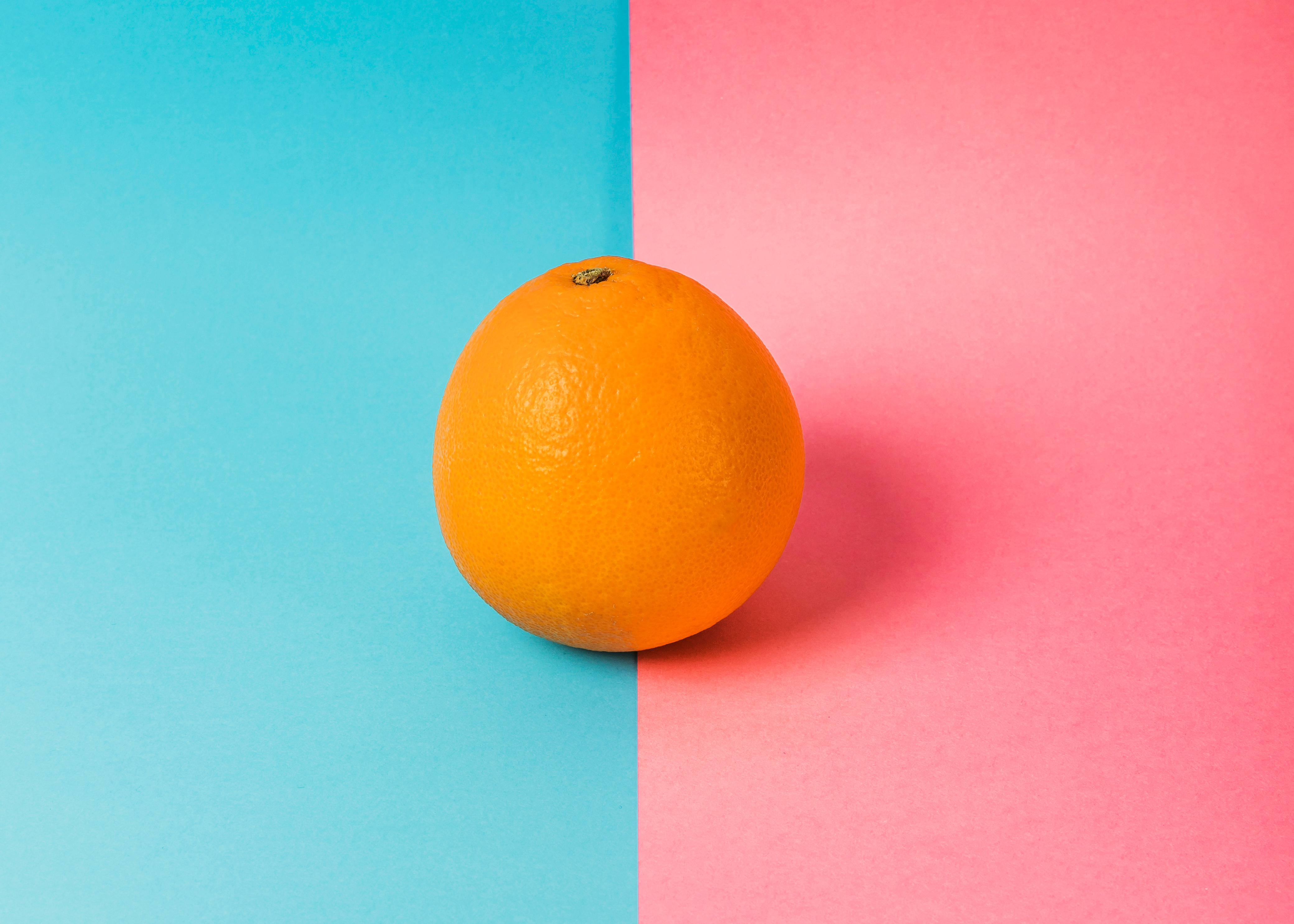 Photo of an orange on a background that if half turquoise and half pink. The orange sits on the dividing line. 